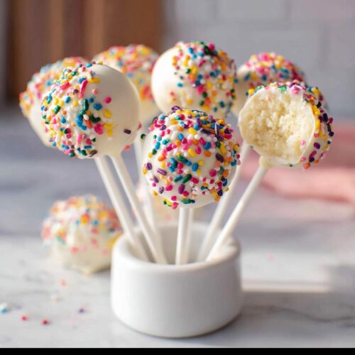 Several White Chocolate Sprinkle Cake Pops dipped in white coating and covered in rainbow sprinkles, displayed in a small white holder.