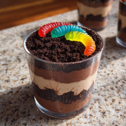 A layered Worms & Dirt Cups dessert in a glass, topped with crushed chocolate cookies and three colorful gummy worms.