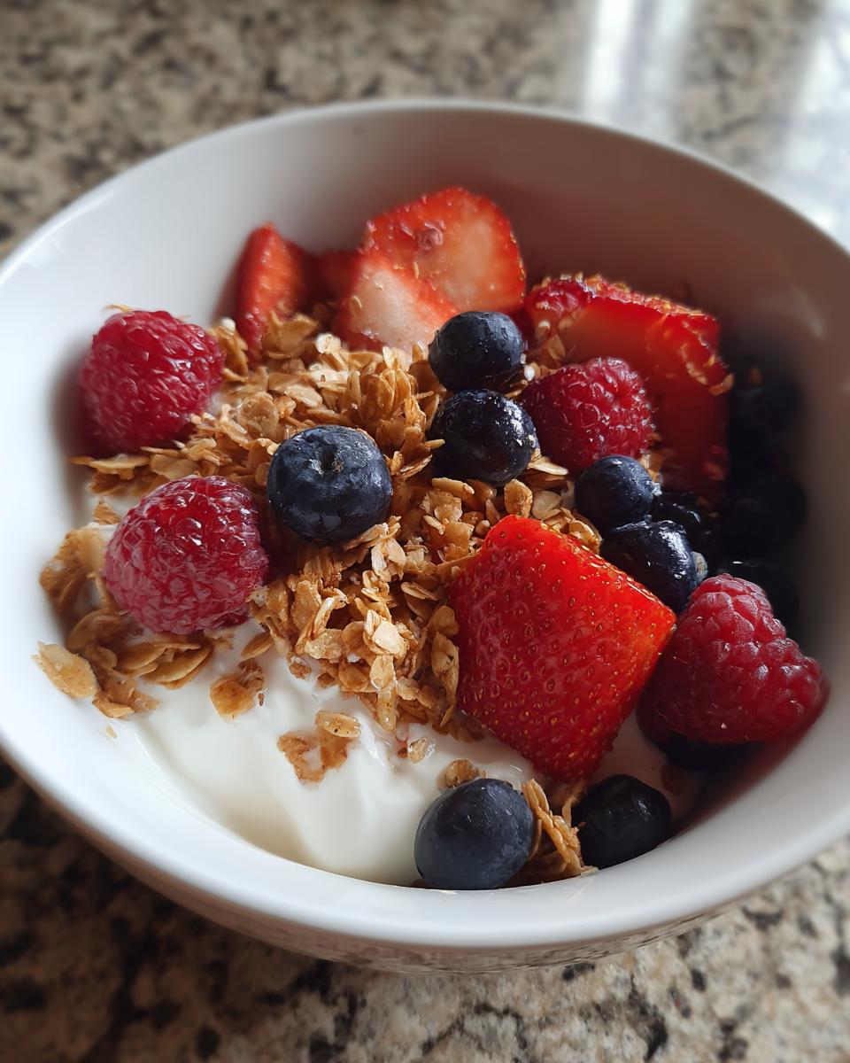 Close-up of a white bowl containing yogurt, granola, blueberries, raspberries, and sliced strawberries, a perfect Quick Workday Breakfast Idea.