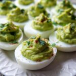 A close-up of several Avocado Deviled Eggs on a white platter, topped with paprika and parsley.