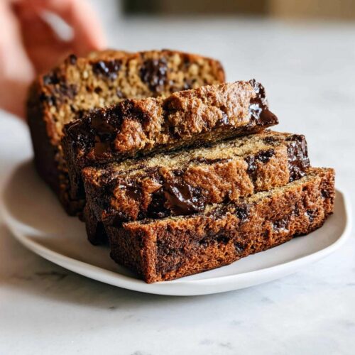 Three thick slices of moist Bakery-Style Chocolate Chip Banana Bread stacked on a white plate, showing melted chocolate chips.