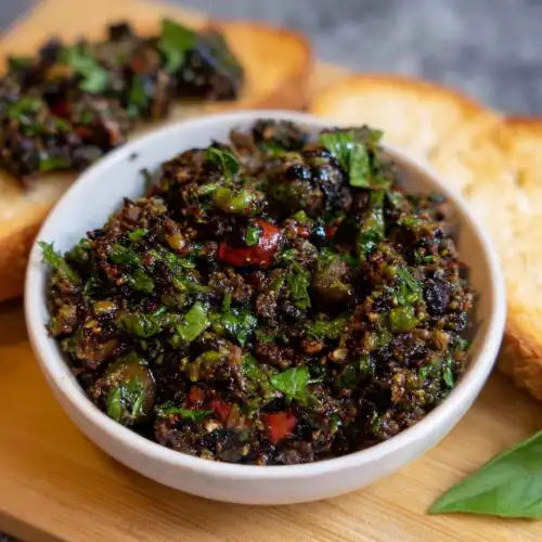 Close-up of a white bowl filled with chunky, dark Olive Tapenade Bruschetta Dip, garnished with fresh green herbs, next to toasted bread.