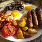 A hearty Brunch Plate Inspired by England featuring sausages, fried eggs, baked beans, roasted tomatoes, potatoes, and mushrooms.