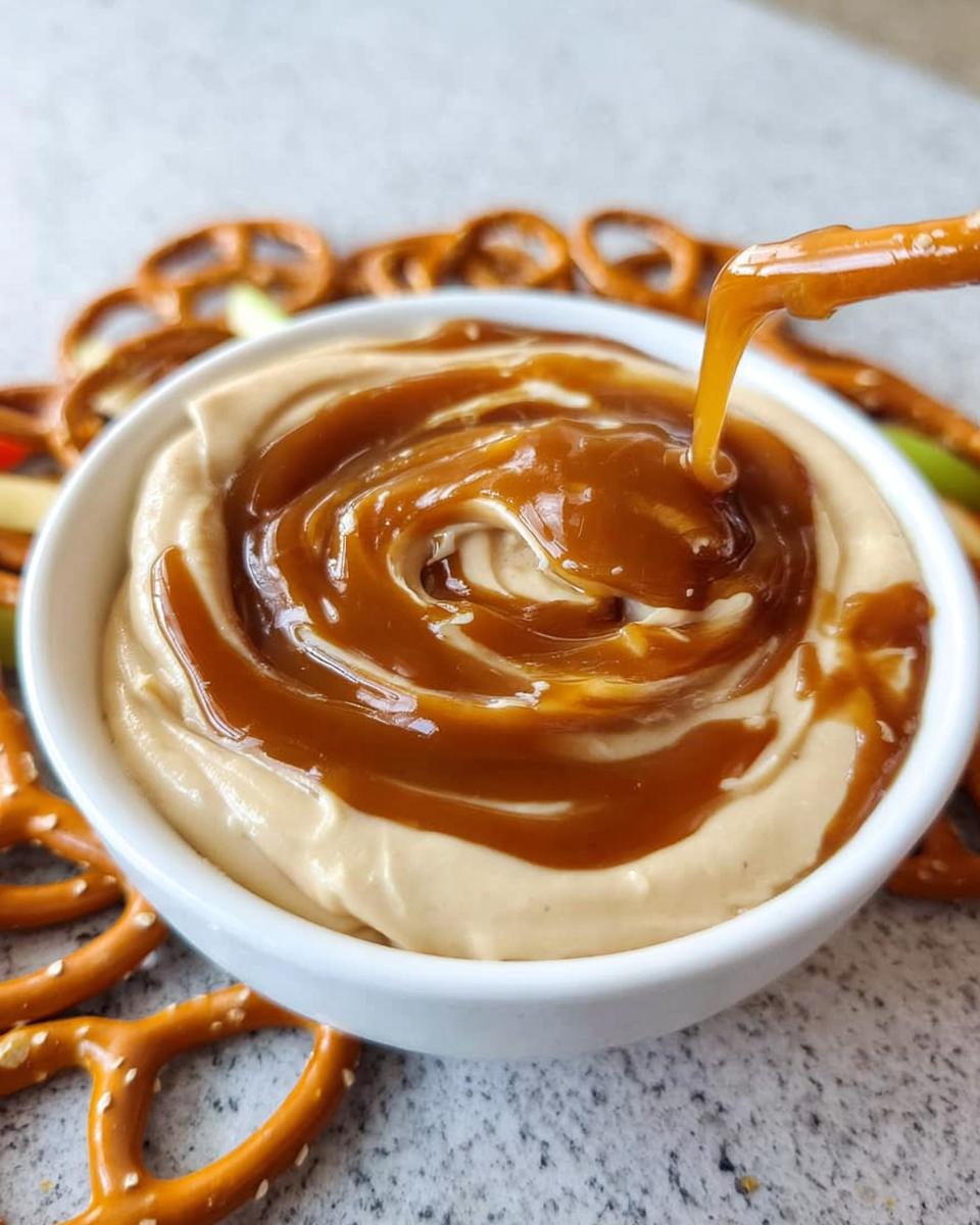 A pretzel being dipped into a bowl of creamy Caramel Pretzel Dip topped with a thick caramel swirl.
