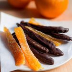 A plate featuring both sugared and dark chocolate-dipped candied orange peel strips, with fresh oranges blurred in the background.