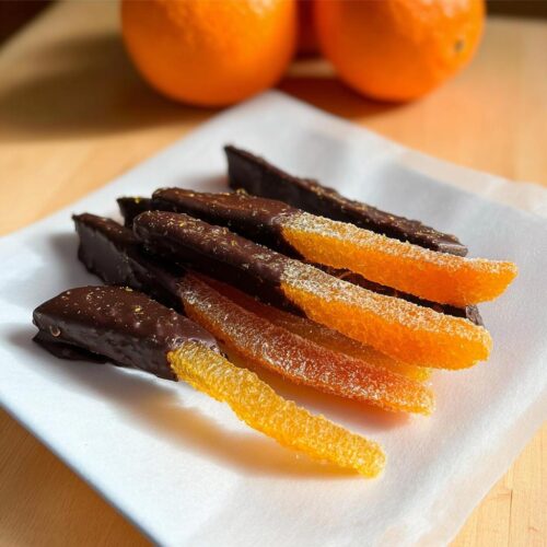 A pile of homemade Chocolate-Dipped Candied Orange Peel strips resting on white parchment paper.