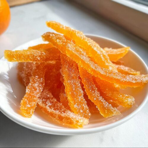 Close-up of bright orange strips of Classic Candied Orange Peel coated in sparkling sugar, piled in a small white bowl.