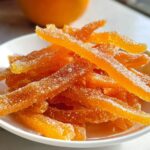 Close-up of bright orange, sugar-coated strips of Classic Candied Orange Peel piled on a small white dish.