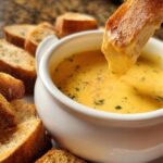 A piece of toasted bread being dipped into a bowl of rich, yellow Classic Garlic Herb Cheddar Fondue.