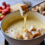 A piece of bread is dipped into bubbling Classic Gruyere Cheese Fondue, creating a long, gooey cheese pull.
