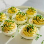 Six beautifully piped Deviled Eggs with Chives and a light sprinkle of paprika on a white platter.