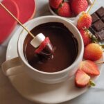 A marshmallow and strawberry dipped in rich Dark Chocolate Fondue served in a white mug.