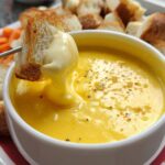 A piece of toasted bread is dipped into a bowl of rich, yellow Garlic Cheddar Cheese Fondue.