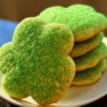 A stack of clover-shaped Green Velvet Sugar Cookies generously covered in bright green sparkling sugar.