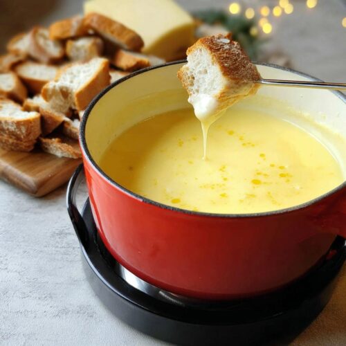 A piece of bread dipped into a red pot of melted Gruyere and Swiss Cheese Fondue, showing a cheese drip.
