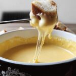 A piece of bread on a fondue fork dripping with melted Gruyere and Emmental Cheese Fondue.