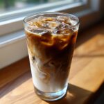 A tall glass of Iced Chai Latte with Oat Milk showing swirling layers of dark chai concentrate and white oat milk over ice cubes.