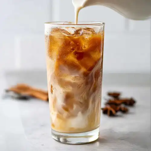 Cream being poured into a tall glass filled with ice and brown liquid for an Iced Chai Latte Made from Tea Bags.