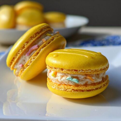 Two bright yellow Lucky Charms Lemon Macarons filled with creamy frosting and colorful cereal pieces, sitting on a white plate.