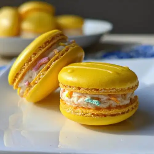 Two bright yellow Lucky Charms Lemon Macarons filled with creamy frosting and colorful cereal pieces, sitting on a white plate.