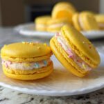 Two bright yellow Lucky Charms Lemon Macarons filled with light pink frosting and colorful sprinkles resting on a white plate.