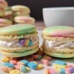 Two colorful Lucky Charms Marshmallow Cream Macarons filled with white cream and colorful cereal pieces.