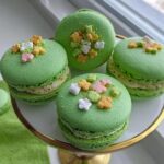 Four vibrant green Lucky Charms Pistachio Macarons topped with colorful marshmallow shapes on a white and gold cake stand.