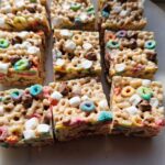 Close-up of square Lucky Charms Rice Krispie Treats with chocolate chips and marshmallows on a white surface.