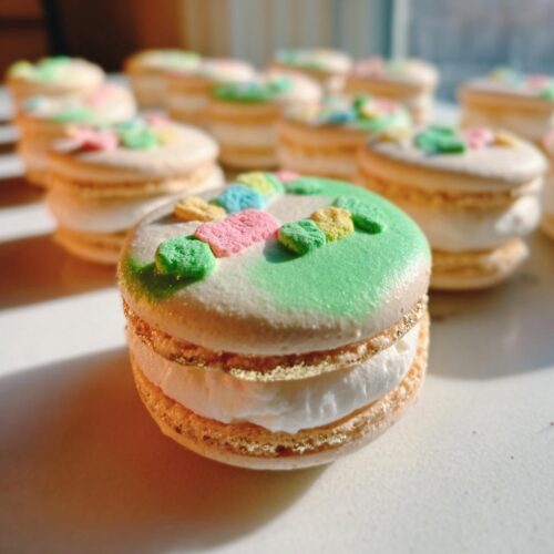 A close-up of a Lucky Charms Vanilla Buttercream Macaron with green icing and colorful marshmallow pieces.