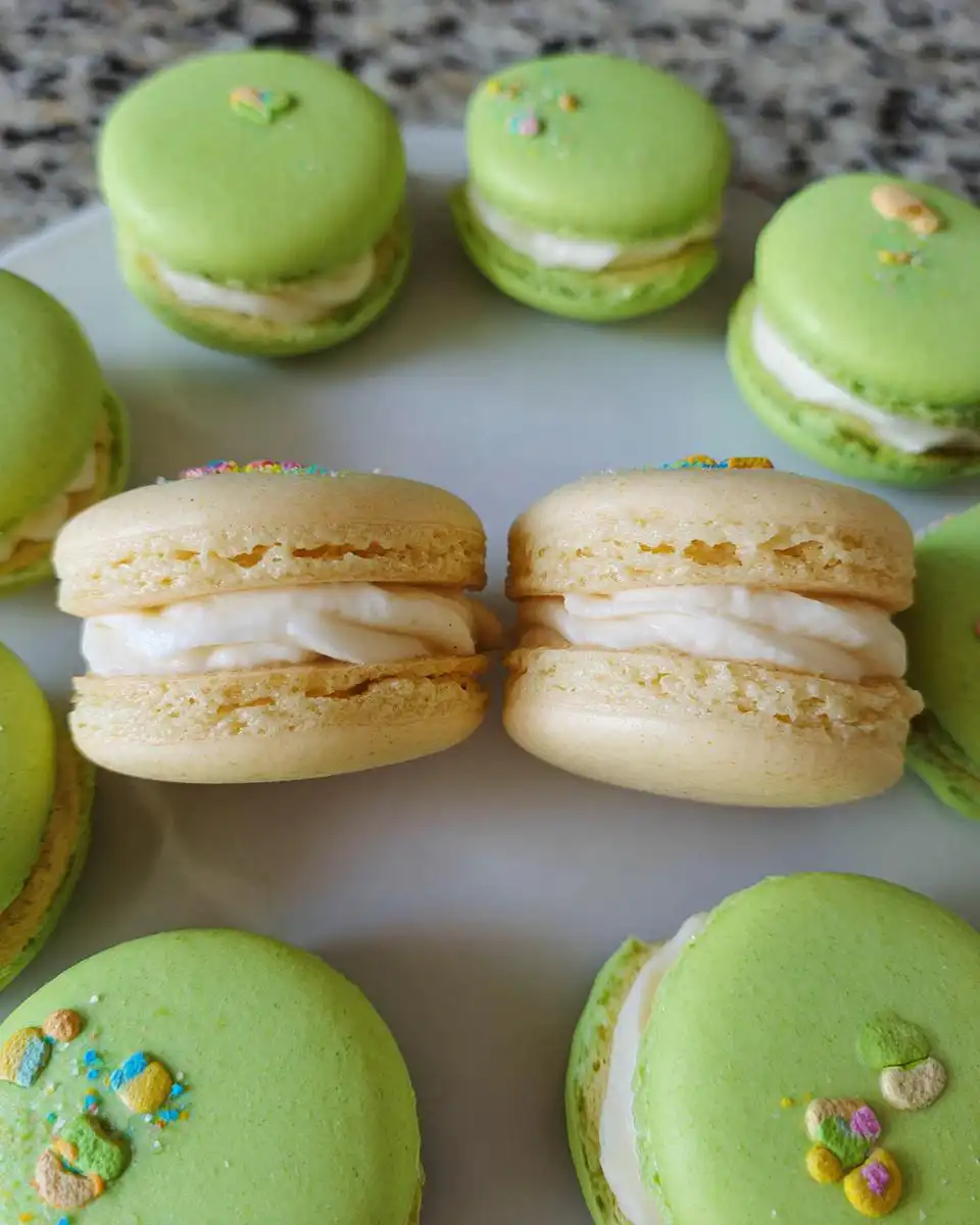 A close-up of several Lucky Charms White Chocolate Macarons, featuring green and tan shells with white filling and colorful sprinkles.