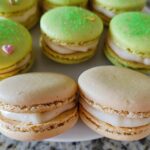 A plate featuring several Lucky Charms White Chocolate Macarons, some green and sprinkled, others plain beige with white filling.