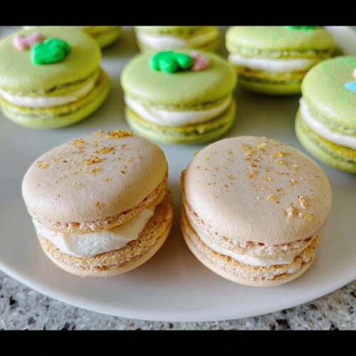 Two beige Lucky Charms White Chocolate Macarons sprinkled with gold dust, served on a white plate with green macarons in the background.