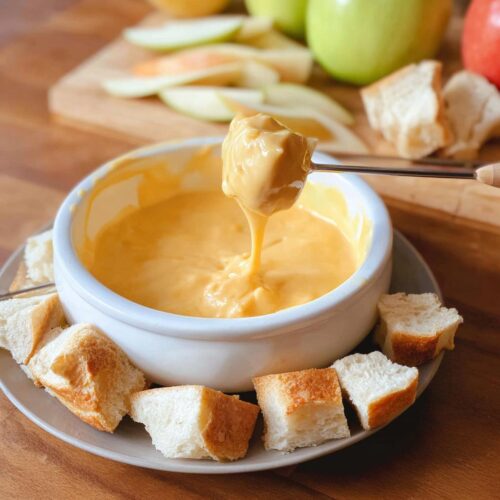 A piece of bread is dipped into a bowl of smooth, yellow Melting Pot Cheddar Cheese Fondue Copycat.