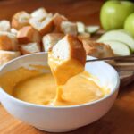 A piece of bread is dipped into a bowl of smooth, bright orange Melting Pot Cheddar Cheese Fondue Copycat.