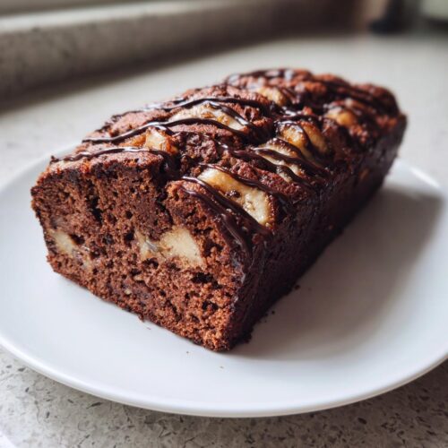 A freshly baked, dark chocolate banana cake loaf topped with sliced bananas and a chocolate drizzle.