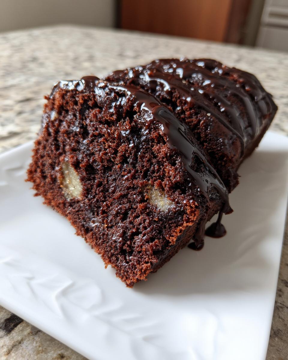 A close-up of a slice of Moist Chocolate Banana Cake, showing the dark crumb and chunks of banana, topped with chocolate glaze.
