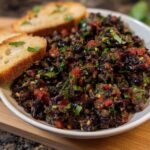 A bowl of chunky Olive Tapenade Bruschetta Dip served with slices of toasted baguette.