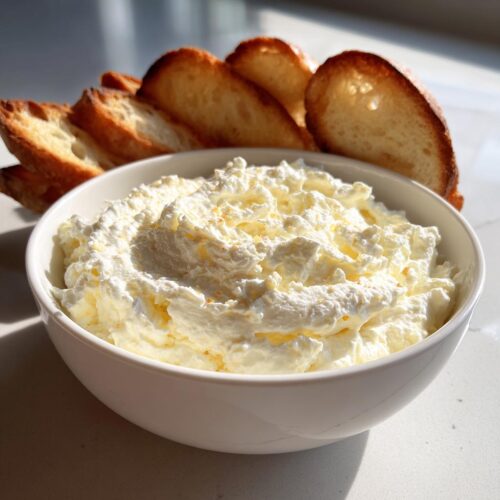 A white bowl filled with creamy Roasted Garlic Bruschetta Dip, served alongside slices of toasted baguette.