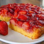 A slice of Simple Strawberry Upside Down Dessert with a whole fresh strawberry beside it on a white plate.