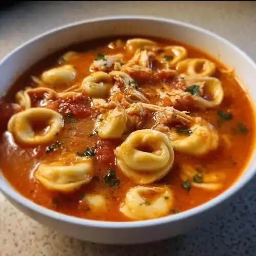 Close-up of a white bowl filled with Slow Cooker Chicken Tortellini in a rich, reddish-orange broth with shredded chicken and parsley.