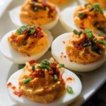 Close-up of several Spicy Sriracha Deviled Eggs topped with chives and red spice on a white platter.