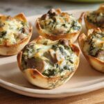 Five baked Spinach Artichoke Mushroom Cups topped with melted cheese and mushrooms, served on a light-colored plate.