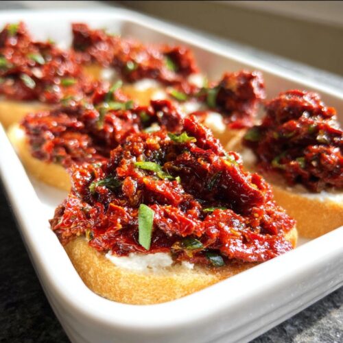 Close-up of small toasts topped with creamy cheese and vibrant Sun-Dried Tomato Bruschetta Dip, garnished with herbs.