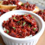 A white ramekin filled with creamy white cheese topped generously with vibrant Sun-Dried Tomato Bruschetta Dip and fresh herbs.