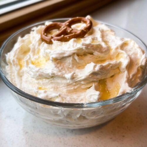 A close-up of Sweet Cream Cheese Pretzel Dip, swirled high in a glass bowl and drizzled with honey, topped with one small pretzel.