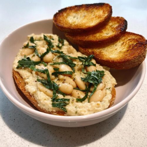 A serving of creamy White Bean Bruschetta Dip spread on toasted bread, topped with whole white beans and fresh basil.