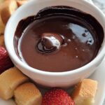 Close-up of smooth, melted chocolate in a white bowl for 3-Ingredient Chocolate Fondue, surrounded by strawberries and pound cake cubes.