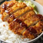 Close-up of a white bowl filled with white rice, topped with sliced, crispy Air Fryer Chicken Katsu drizzled with brown sauce.