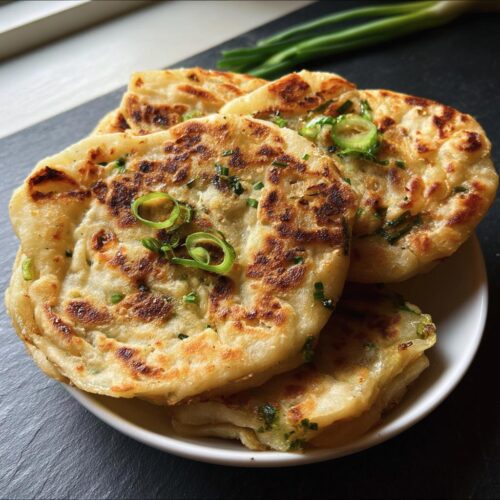 A stack of golden-brown Air Fryer Scallion Pancakes garnished with fresh green onion slices on a white plate.