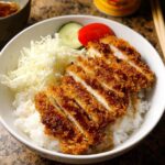 A white bowl filled with white rice topped with sliced, crispy Baked Chicken Katsu Bowl, shredded cabbage, and a tomato slice.